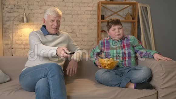 爷爷和孙子坐在沙发上看电视吃着薯片微笑着一位老人视频的预览图