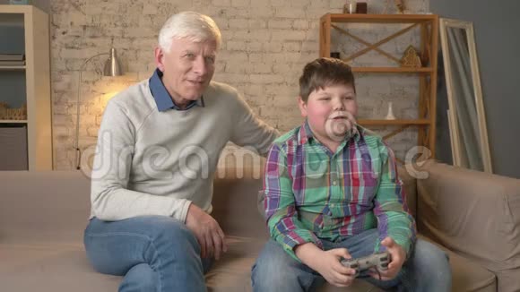 爷爷看着孙子玩游戏机老人正和一个小胖子坐在沙发上视频的预览图
