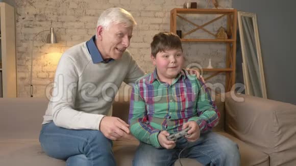 爷爷在控制台游戏中享受孙子的胜利老人正和一个小胖子坐在沙发上视频的预览图
