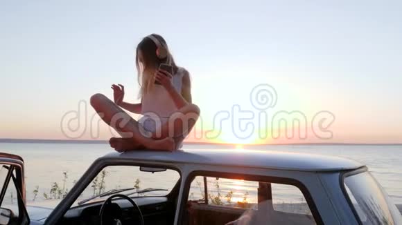 夏天在汽车上旅行女孩在车顶上听耳机音乐女孩在汽车上欣赏日落时的好音乐快乐视频的预览图