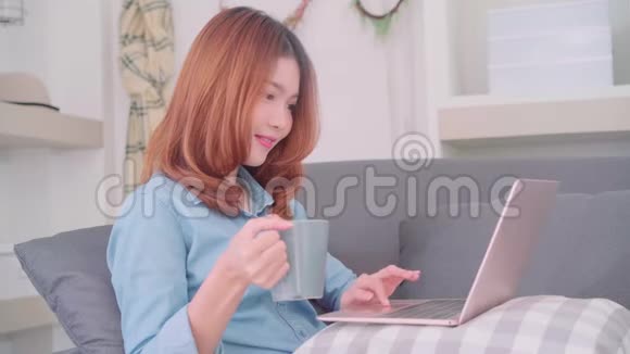 一幅美丽迷人的亚洲女人在沙发上躺着时用电脑或笔记本电脑拿着一杯温暖的咖啡或茶的肖像视频的预览图
