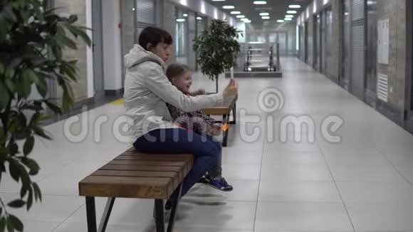 母亲和女儿在商场或办公室的智能手机上自拍视频的预览图