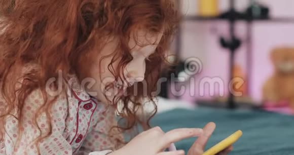 有趣的红头发小女孩与黄色智能手机躺在床上和使用智能手机沟通播放APP视频的预览图
