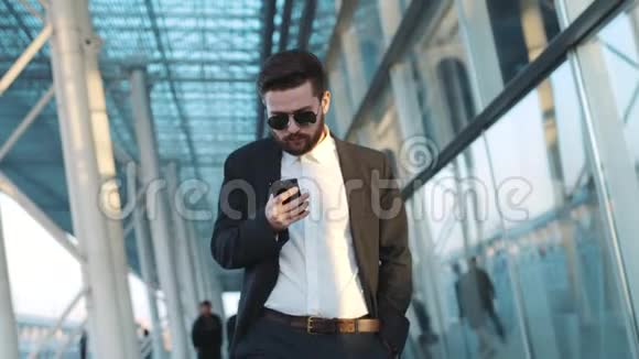 年轻的大胡子男子走过机场候机楼在手机上注意到smth惊讶地摘下太阳镜视频的预览图