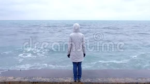 穿着白色夹克的女人站在海滩上的海滨看着暴风雨的波浪后景视频的预览图