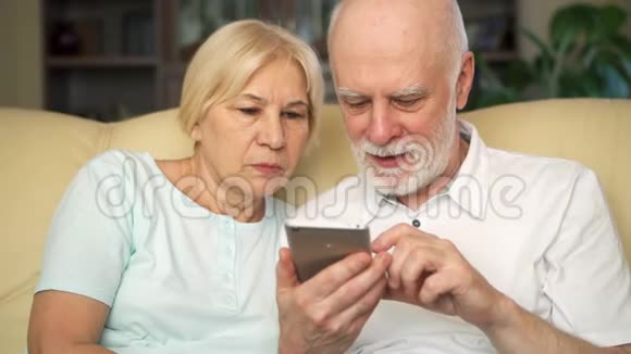 老两口坐在家里的沙发上现代养老金领取者使用移动浏览阅读新闻视频的预览图