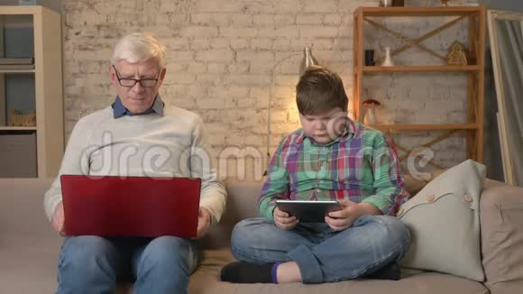 代的差异戴眼镜的老人坐在沙发上用笔记本电脑工作年轻的胖男孩在玩视频的预览图