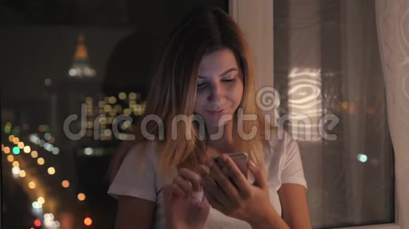 夜晚的女人站在窗口使用智能手机视频的预览图