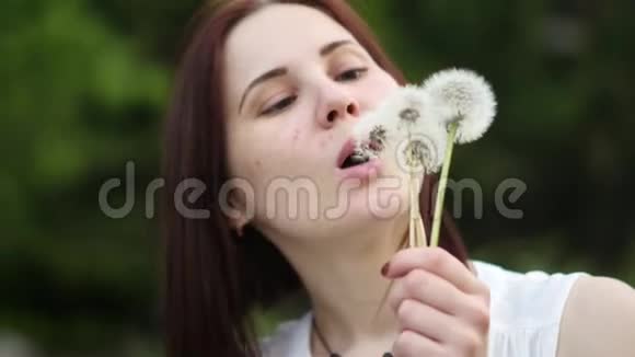 一个年轻可爱的女人在一个夏日绿色花园的背景下吹着蒲公英视频的预览图