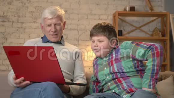 祖父和孙子坐在沙发上在笔记本电脑上看着一部有趣的电影笑着家庭视频的预览图