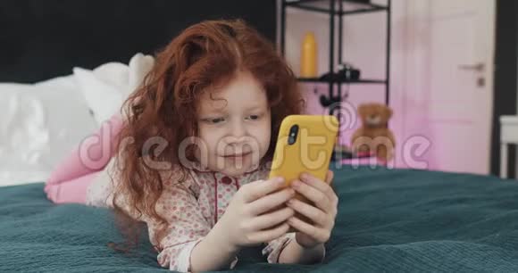 有趣的红头发小女孩与黄色智能手机躺在床上和使用智能手机沟通播放APP视频的预览图