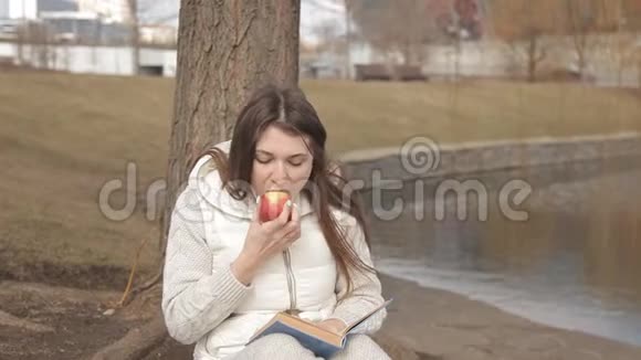 美丽的女孩在公园里的一棵树下吃一个红苹果在水边看书视频的预览图