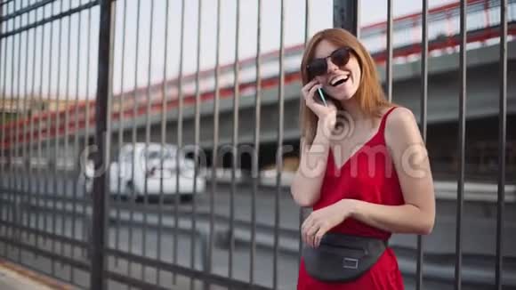 街上漂亮的红发年轻女子的肖像穿着红色连衣裙使用智能手机慢动作视频的预览图