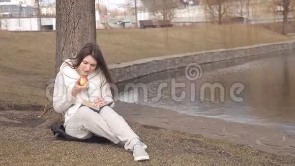 美丽的女孩在公园里的一棵树下吃一个红苹果在水边看书视频的预览图