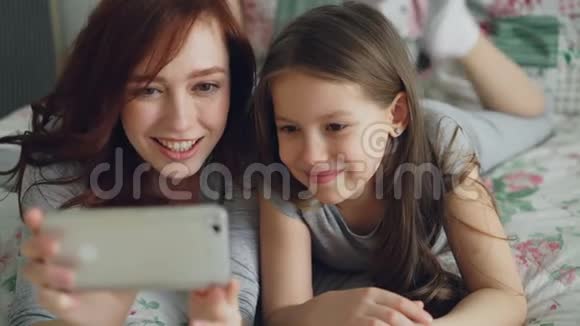 特写快乐妈妈和小女孩用智能手机相机自拍坐着玩开心的鬼脸视频的预览图