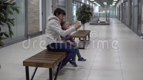 母亲和女儿在商场或办公室的智能手机上自拍视频的预览图