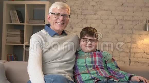 祖父和孙子戴着眼镜坐在沙发上微笑着看着相机家庭舒适家庭舒适视频的预览图