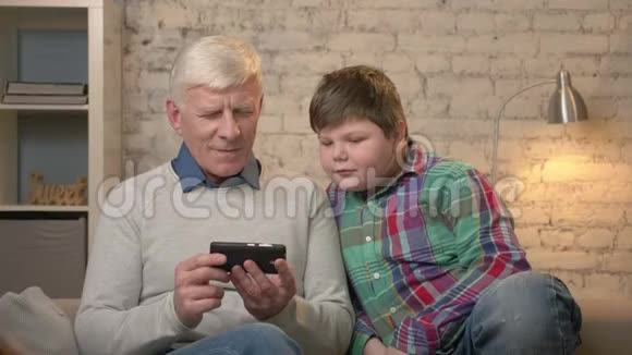 祖父和孙子坐在沙发上用智能手机玩小胖子视频的预览图