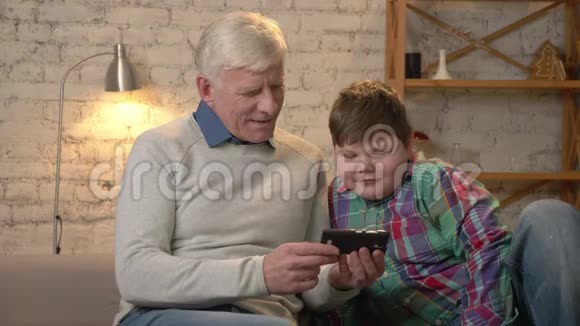 祖父和孙子坐在沙发上用智能手机看有趣的视频有兴趣严肃的脸视频的预览图