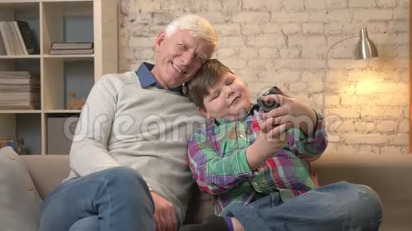 爷爷和孙子坐在沙发上用智能手机做自拍年轻的胖孩子和爷爷微笑视频的预览图