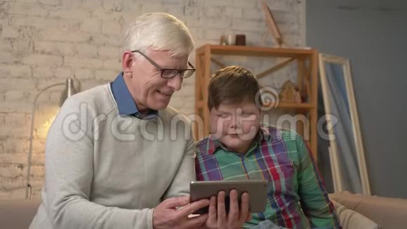 祖父和孙子坐在沙发上用平板电脑看有趣的视频有兴趣严肃的脸视频的预览图