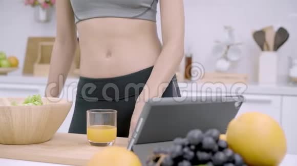 年轻的亚洲妇女做沙拉健康的食物同时使用平板电脑寻找食谱并在厨房喝橙汁视频的预览图