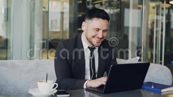 快乐的白种人企业家穿着正式的衣服一边在笔记本电脑上打字一边听音乐耳朵里有耳塞视频的预览图