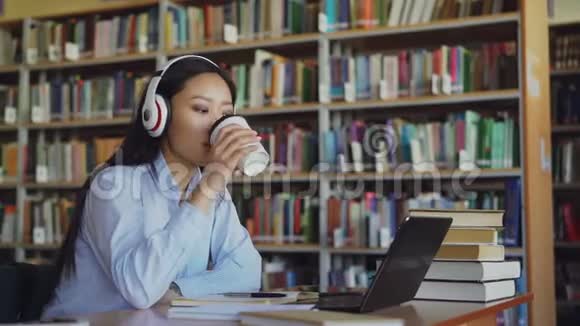 美丽的亚洲女学生坐在图书馆的办公桌前听着耳机里的音乐在学校项目工作青年视频的预览图