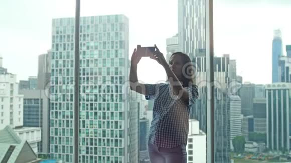 快乐的黑发女人在漂亮的城市视野的公寓靠窗用手机自拍照片3840x2160视频的预览图