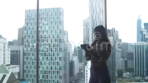 美丽的年轻女子站在窗边在城市的背景下使用智能手机3840x2160视频的预览图