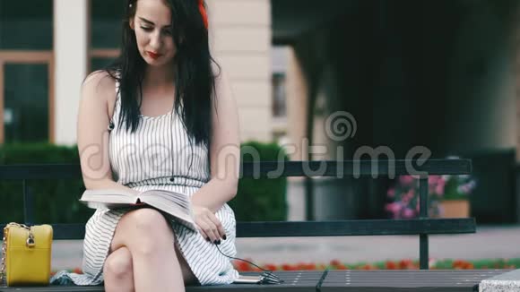 一个头上有红色边缘的漂亮女孩坐在公园的长凳上看书然后拿着手机视频的预览图