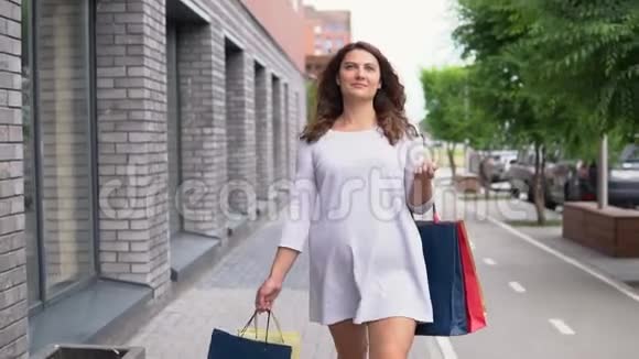 一个穿着浅色衣服的女孩购物后心情愉快地走在街上带着购物的包裹视频的预览图