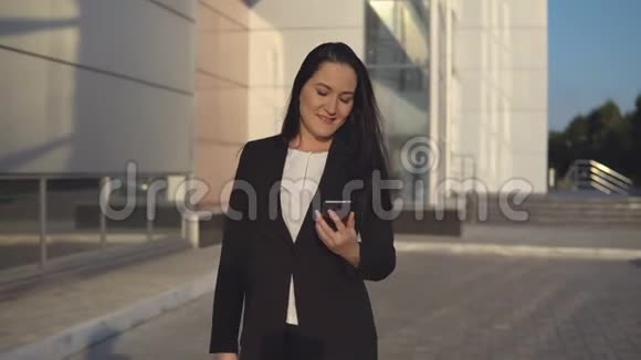 穿着黑色西装的年轻女孩在办公室中心户外的背景下在智能手机上交谈视频的预览图