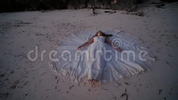 夕阳西下一位美丽而快乐的新娘躺在沙滩上在她周围铺开一件婚纱最初的想法视频的预览图