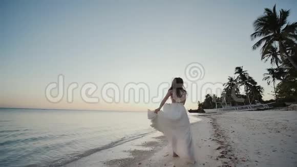 迷人而快乐的新娘穿着白色连衣裙赤脚在热带海滩的沙滩上跳舞海洋手掌视频的预览图