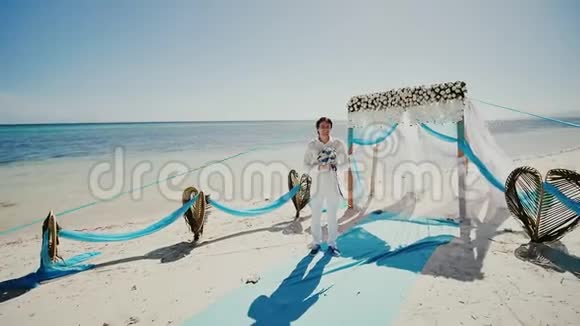 在棕榈树和海洋之间的热带海滩上举行婚礼一个时髦的快乐新郎带着一束白色的花视频的预览图
