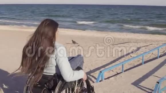 在春季或秋季坐轮椅的残疾青年妇女在靠近海洋的斜坡旁为残疾人移动视频的预览图