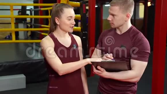 肌肉教练在剪贴板上写字在交叉训练体育馆和年轻女子交谈视频的预览图