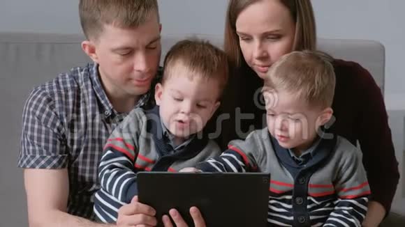 带平板电脑的家人妈妈爸爸和两个儿子双胞胎坐在沙发上玩平板电脑游戏视频的预览图