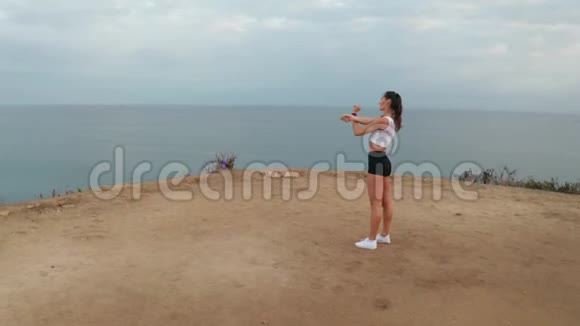 日出时在海边悬崖上伸展的女性鸟瞰图视频的预览图