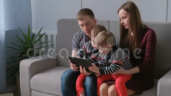带平板电脑的家人妈妈爸爸和两个儿子双胞胎坐在沙发上玩平板电脑游戏视频的预览图