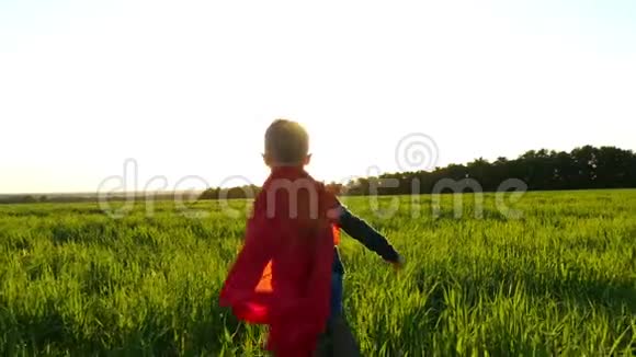 一个穿着超级英雄服装的快乐孩子穿着红色雨衣在日落的背景下沿着绿色的草坪奔跑视频的预览图