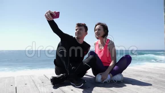 年轻的健康夫妇坐在海滩附近拿着电话摆出自拍姿势快乐的男朋友和女友拍照视频的预览图