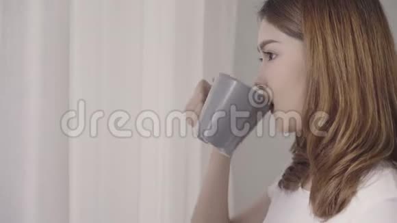 快乐美丽的亚洲女人微笑着在卧室的窗户附近喝一杯咖啡或茶视频的预览图