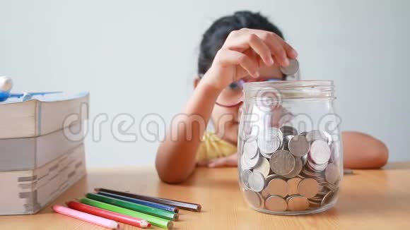 亚洲小女孩把硬币放进一个透明的玻璃瓶桌子上放着一本书比喻为教育理念省钱视频的预览图