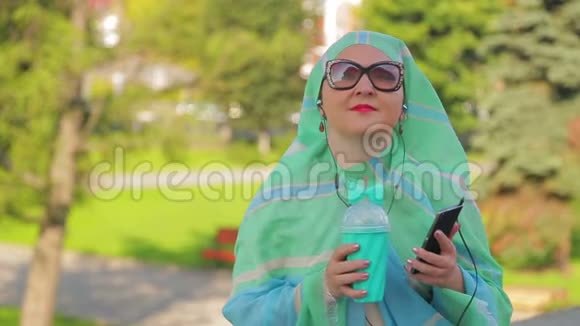 公园里一位戴着浅色围巾的年轻穆斯林妇女正在喝咖啡用耳机在信使里说话视频的预览图