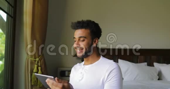 年轻的西班牙裔男子使用平板电脑快乐的微笑男人在网上聊天的大窗口与热带花园景观视频的预览图