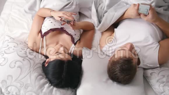 年轻漂亮的情侣躺在床上手里拿着智能手机一起休息每个人都看着你的手机视频的预览图