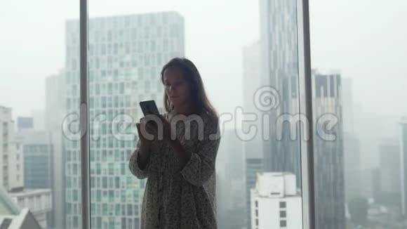 年轻的女性自由职业者或商人使用手机并在豪华公寓的全景窗口交谈视频的预览图