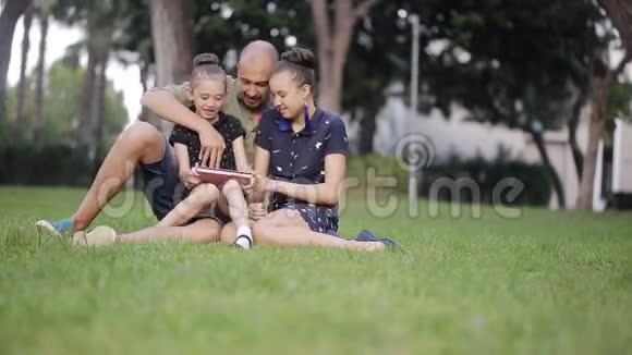 父亲带着两个女儿在草地上从平板电脑屏幕上读到故事笑了起来视频的预览图